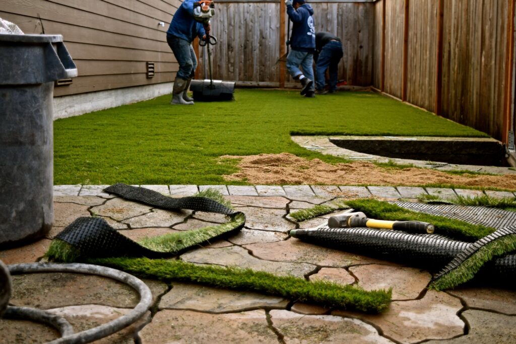 Workers installing artificial grass in a backyard with a wooden fence.