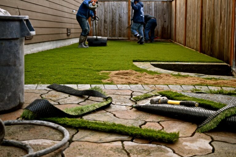 Workers installing artificial grass in a backyard with a wooden fence.