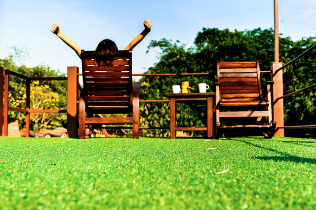 Person relaxing on artificial grass with outdoor furniture and green trees.