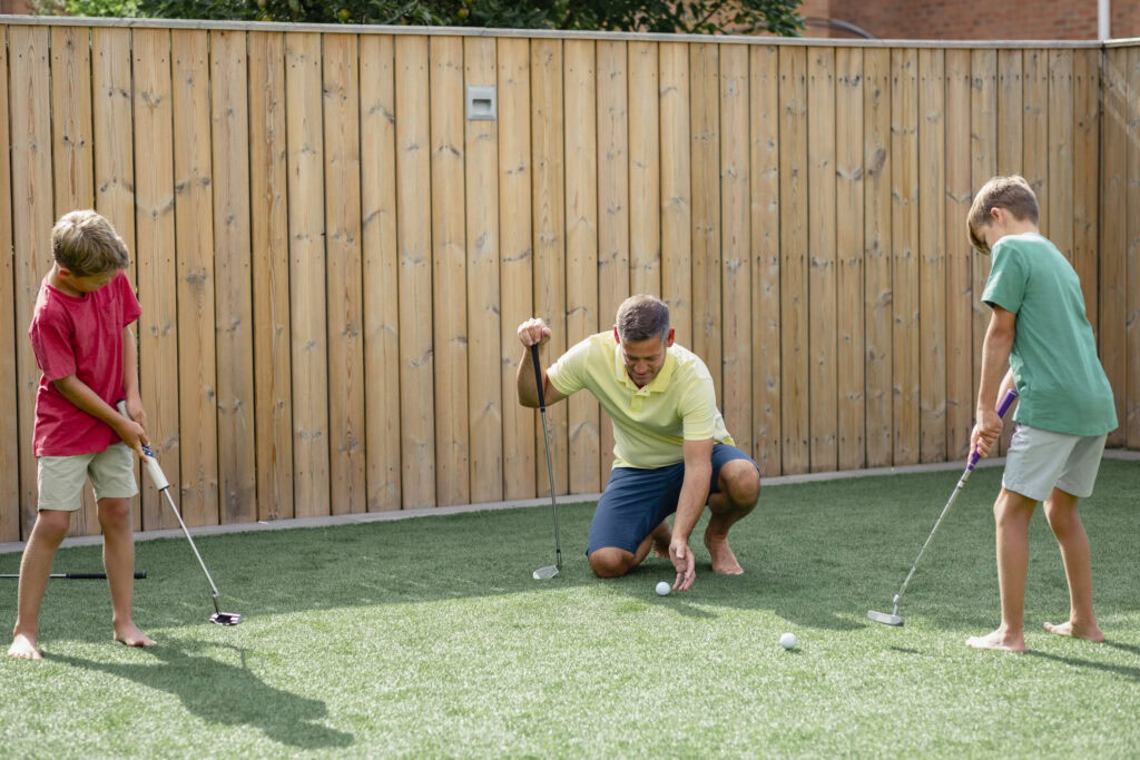 Family playing golf on artificial turf in backyard with wooden fence.