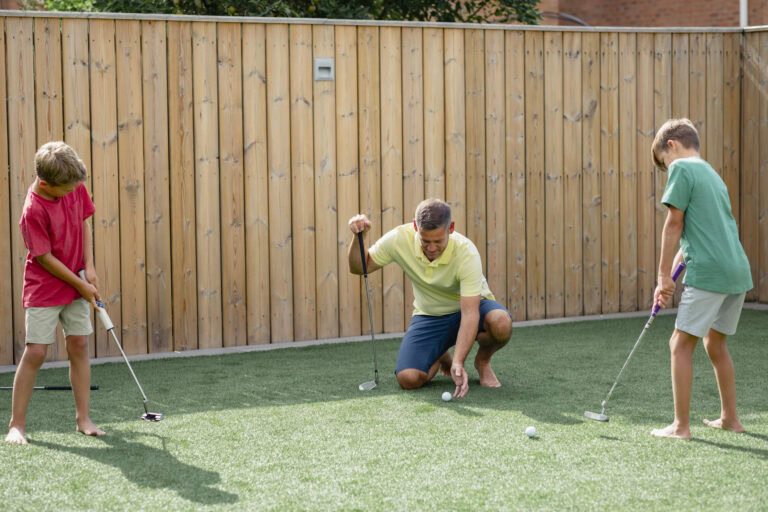 Family playing golf on artificial turf in backyard with wooden fence.