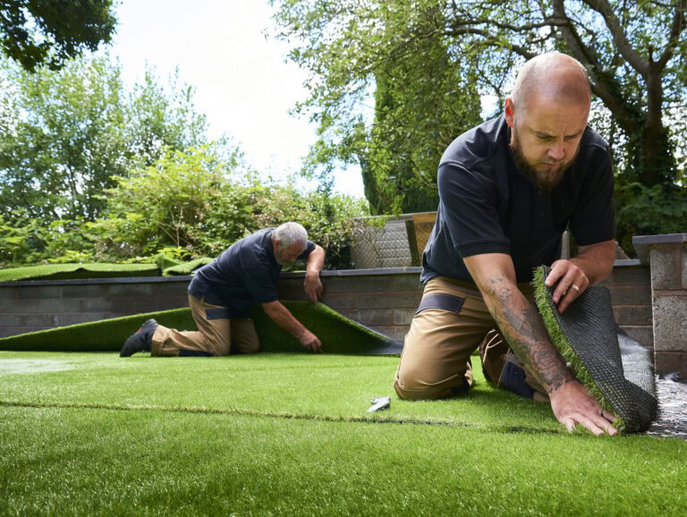 Two workers installing artificial turf in a lush backyard garden.