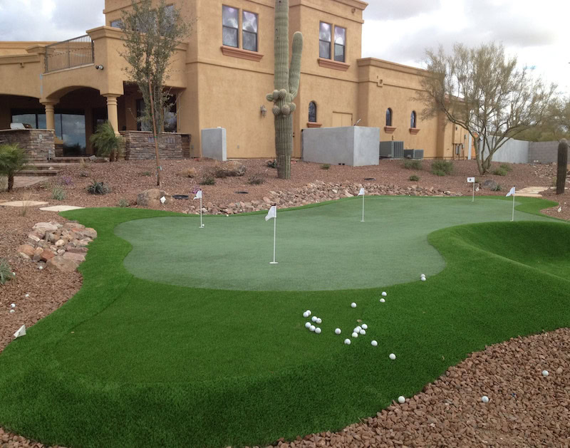 Multi-hole backyard putting green with golf balls and white flag sticks, luxury stucco home with towering saguaro cactus, Scottsdale Arizona