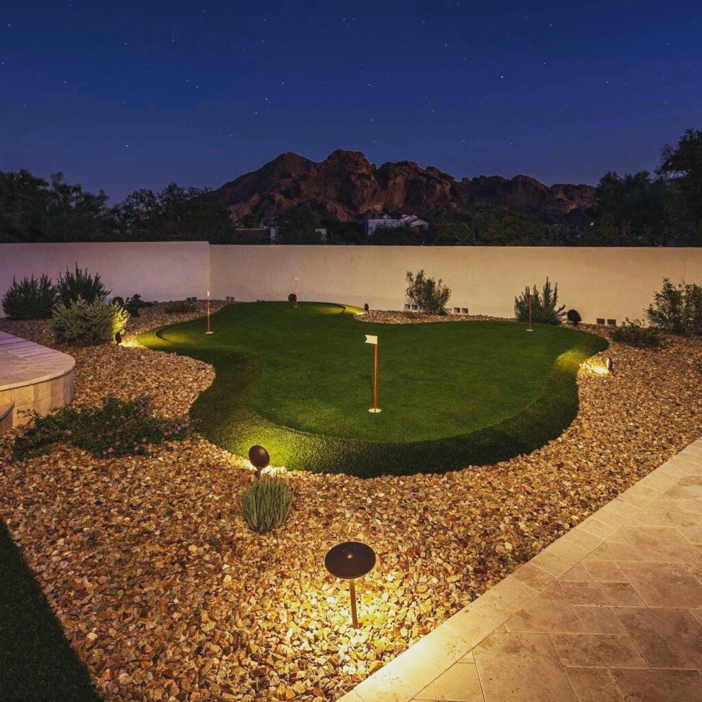 Backyard putting green illuminated by landscape lighting at night with dramatic Camelback Mountain rock formation visible against deep blue sky, Paradise Valley Arizona