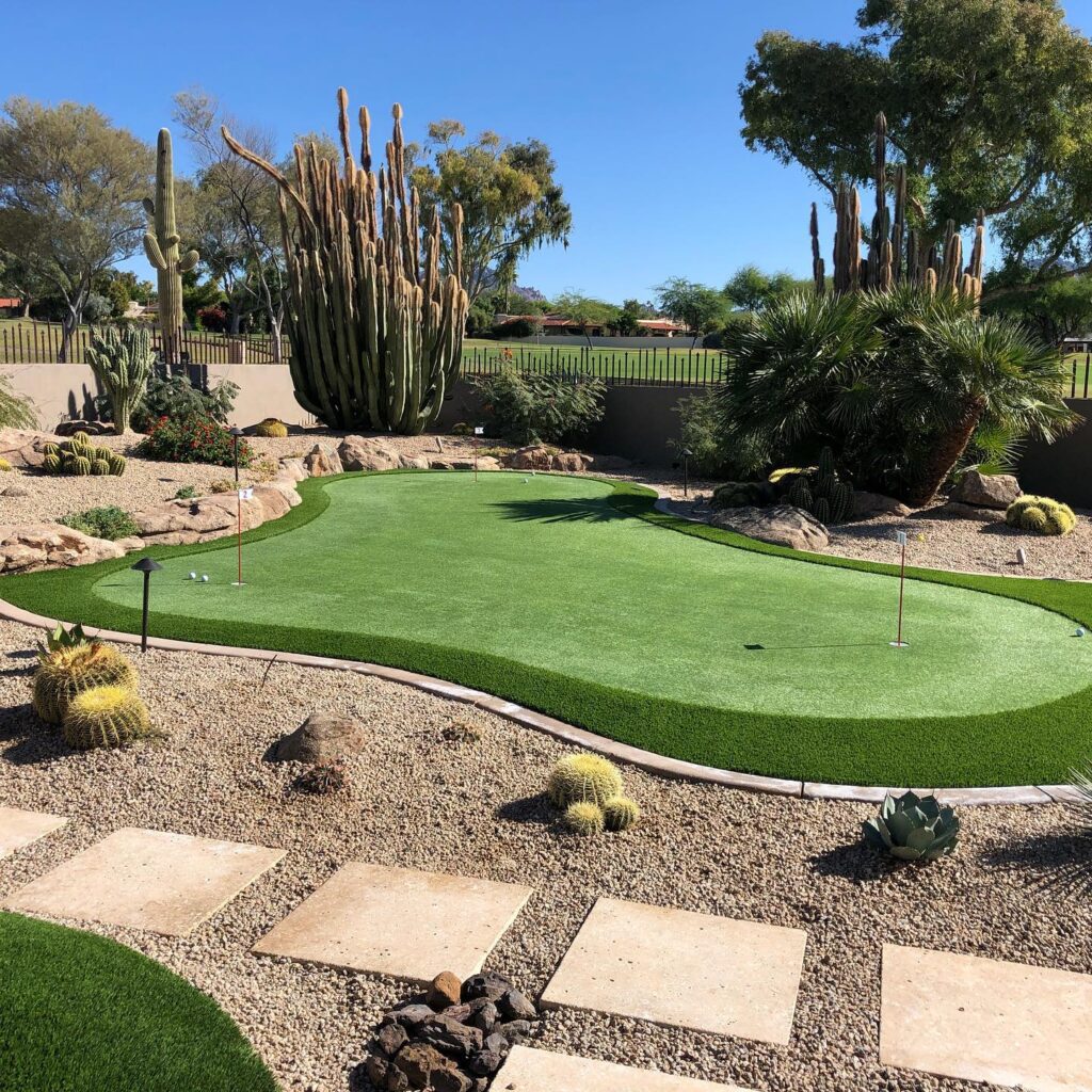Backyard putting green with saguaro and barrel cactus, desert boulders, travertine stepping stones, and golf course visible through iron fence, Scottsdale Arizona