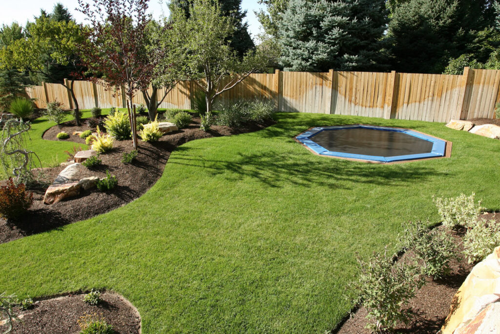 In-ground octagonal trampoline with blue safety cover, surrounded by lush backyard with mulch landscape beds, colorful shrubs, and wood fence