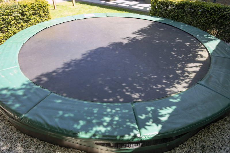 Close-up of round in-ground trampoline with green safety padding flush with surrounding gravel and manicured garden