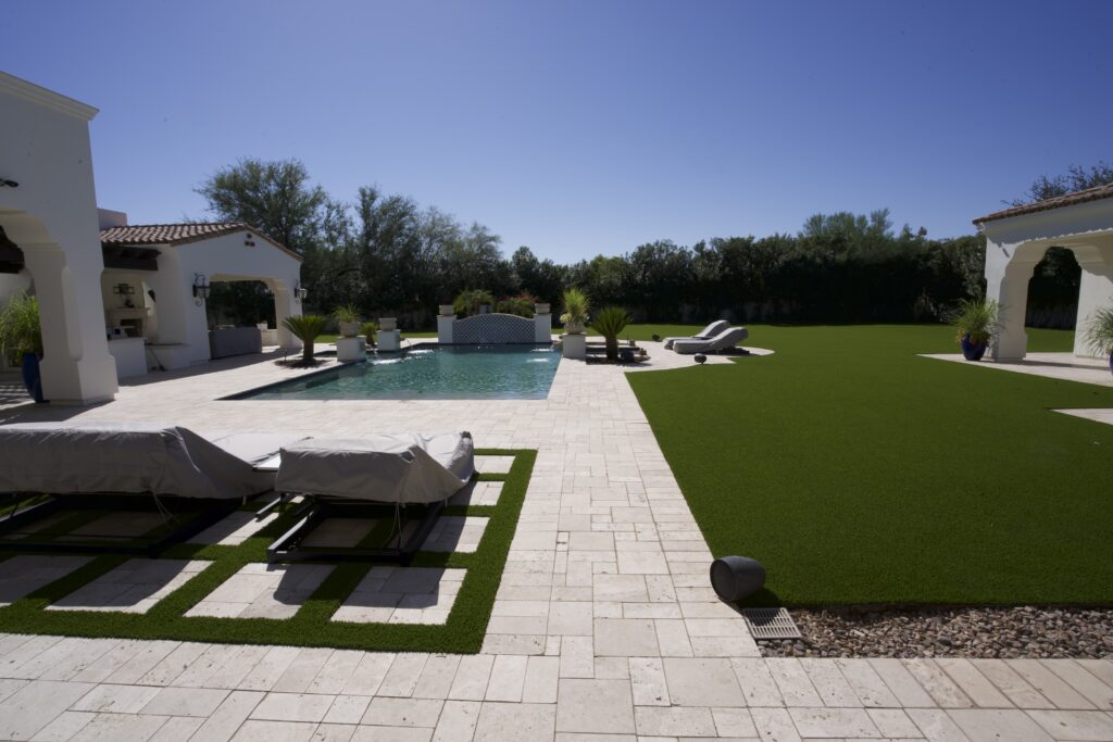 Full backyard view of Scottsdale luxury estate with artificial grass lawn, travertine pool deck, and white stucco home